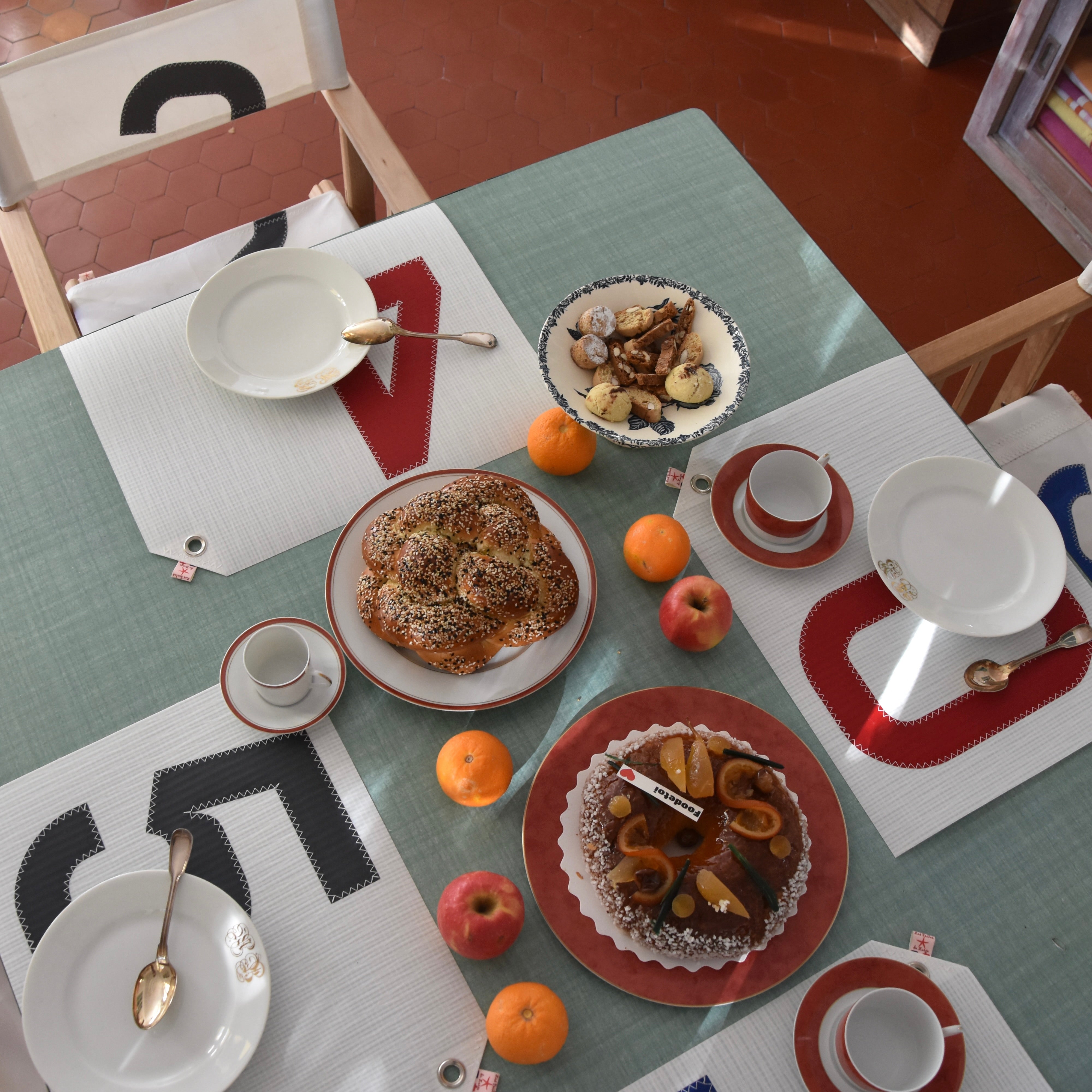 Table de petit-déjeuner vue du dessus, dressée avec des sets de table en voile de bateau recyclée aux motifs graphiques, assiettes et tasses en céramique, brioche, gâteau, fruits frais et viennoiseries, dans une ambiance chaleureuse et conviviale.