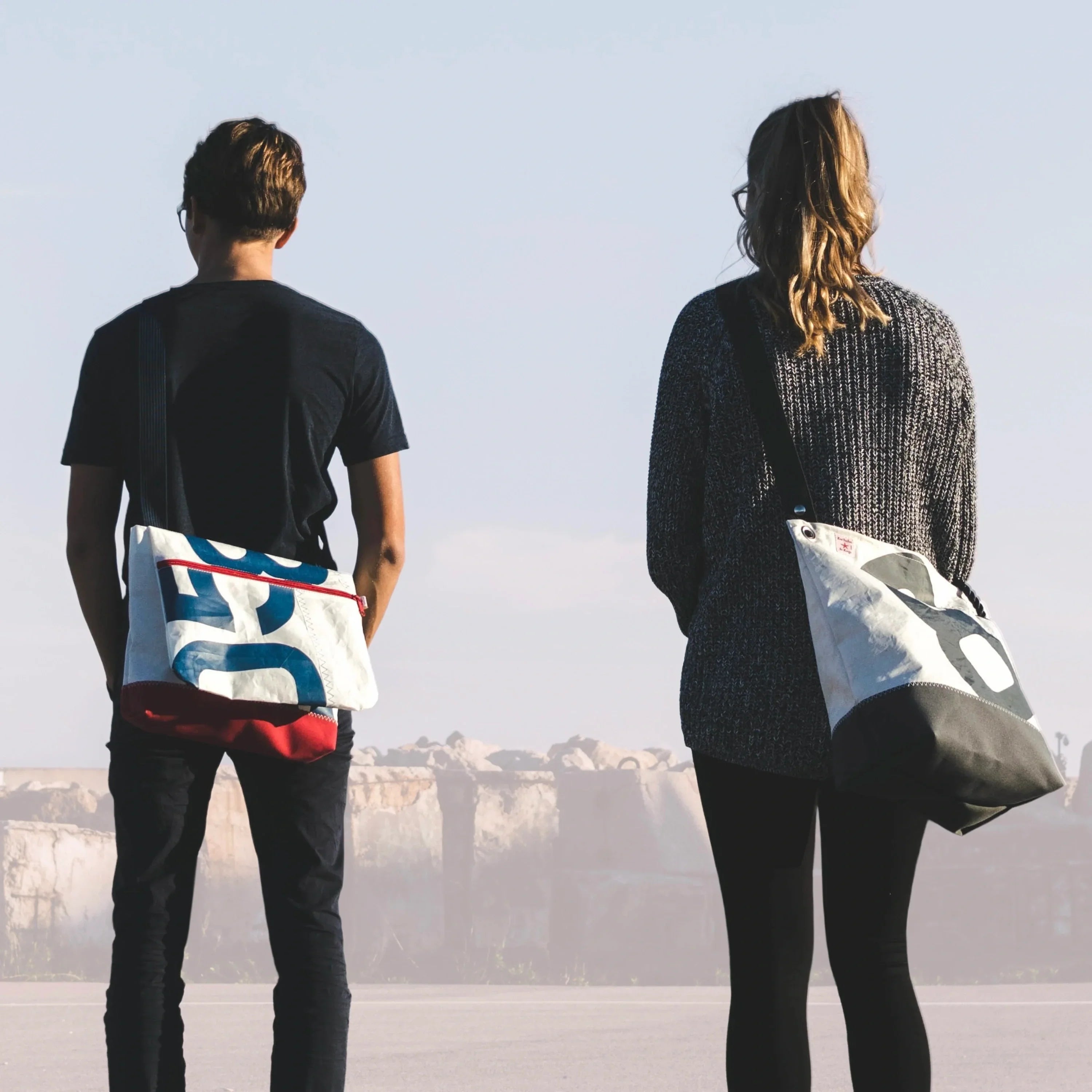 Un jeune homme et une jeune femme de dos. Chacun porte un sac en bandoulière Les Toiles du Large. Ces sacs sont fabriqués à partir de voiles de bateau recyclées à La Ciotat. Ils sont fabriqués de façon artisanal dans un atelier situé sur le vieux-port.